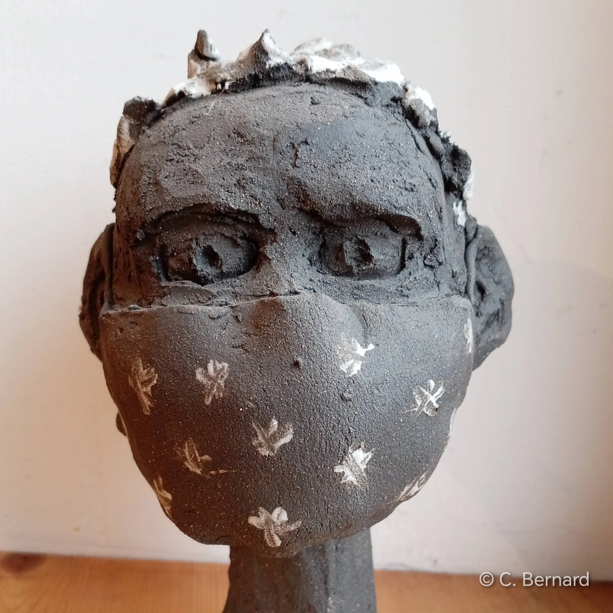 Un visage de grès se dresse, muet et figé, comme pétrifié par l'invisible. Le masque chirurgical y creuse des sillons de solitude et de résilience.